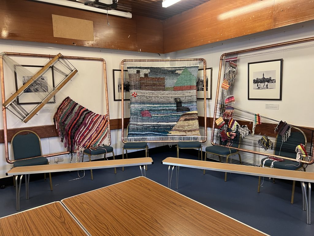 Three different weavings displayed on metal pipes. One is a picture of a seal playing in the sea, with The Pyramid in the background.